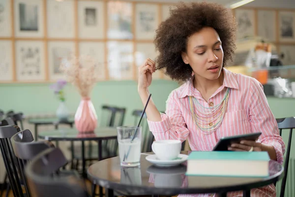 Atrakcyjna kobieta rasy mieszanej w paski różowy strój siedzi w kawiarni i za pomocą tabletu. — Zdjęcie stockowe