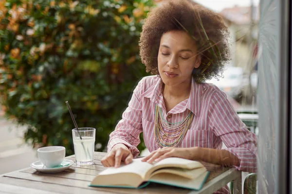 Aantrekkelijke gemengde race vrouw in roze gestreepte jurk lezen boek in café. Op tafel zijn koffie en limonade. — Stockfoto