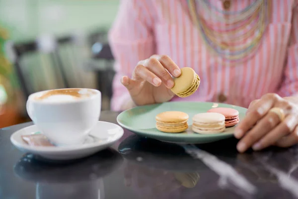 Mujer de raza mixta en vestido de rayas rosadas sosteniendo la galleta mientras está sentado en la pastelería. En la mesa hay café y plato con galletas . — Foto de Stock