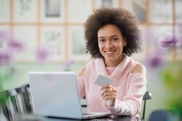 Vacker blandad ras kvinna i rosa polotröja sitter i konditoriet och använder laptop för on line shopping. — Stockfoto
