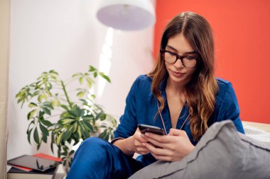 Beyaz esmer büyüleyici mavi pijama ve bedrooma yatakta oturup ve akıllı telefon kullanarak kameraya bakarak gözlük ile giyinmiş. Sabah saat.