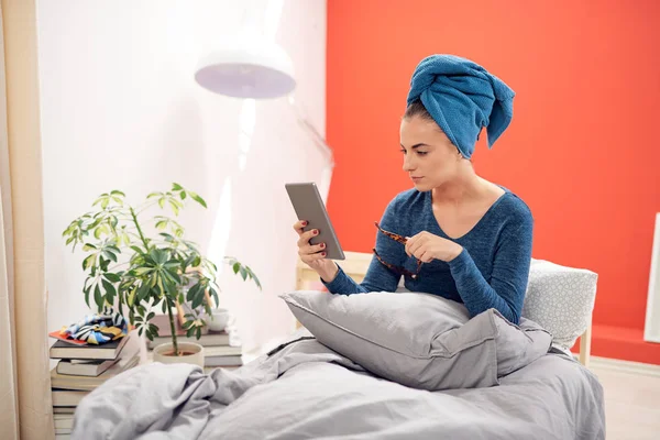 Hermosa morena con toalla en la cabeza usando tableta mientras está sentada en la cama en el dormitorio . — Foto de Stock