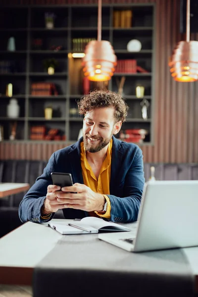 Ung vit affärsman klädd smart casual med hjälp av smart telefon medan du sitter på café. På skrivbordet är laptop. — Stockfoto