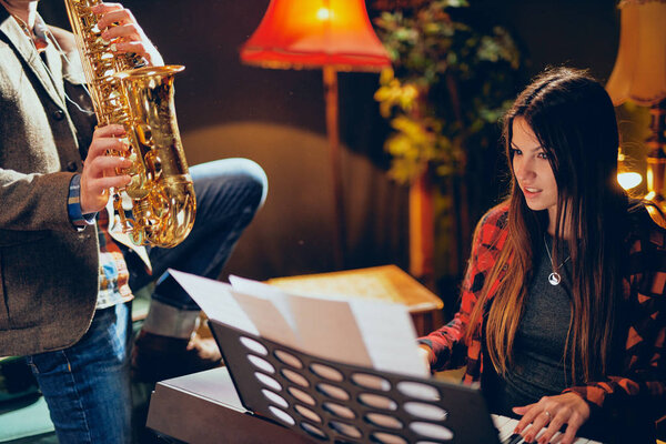 Caucasian man playing saxophone in home studio while young talented woman playing clavier.