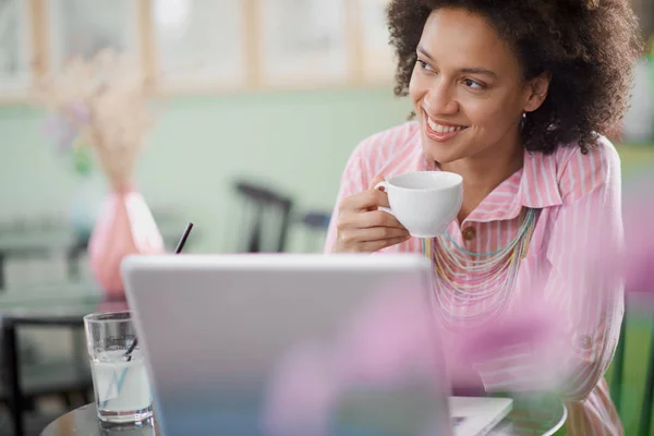 Encantadora mujer de raza mixta en vestido rosa a rayas sentado en la cafetería, beber café y usar el ordenador portátil . —  Fotos de Stock