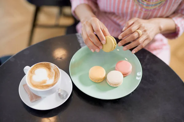 Top view of mixed race woman in pink striped dress holding cookie while sitting in pastry shop. On table are coffee and plate with cookies. — Stock Photo, Image