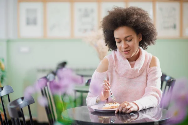 Portret van charmante positieve mixed race vrouw in roze coltrui zitten in zoetwaren en eten gateau. — Stockfoto