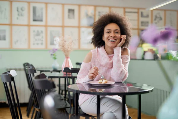Porträt einer charmanten positiven Mischlingsfrau im rosafarbenen Rollkragenpullover, die in einer Konditorei sitzt und Torte isst. — Stockfoto
