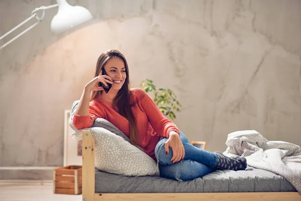 Joven morena caucásica sentada en la cama con auriculares en las orejas y usando un teléfono inteligente. Fondo rojo coral . — Foto de Stock