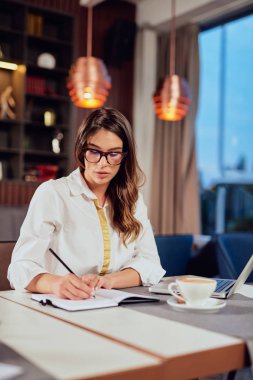 Young beautiful serious Caucasian brunette sitting at cafe and writing in agenda tasks. Remote business concept.