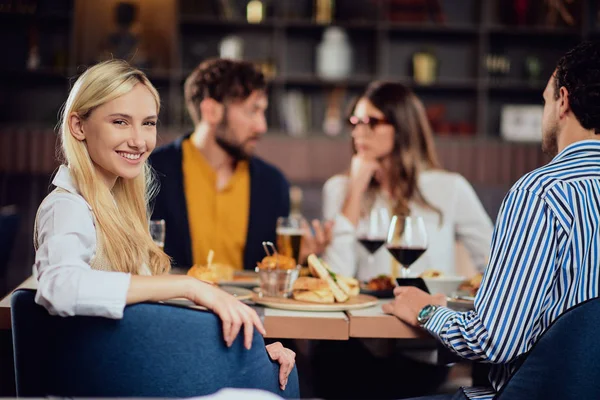 Jonge glimlachende blanke blonde leuke vrouw gekleed slimme casual zitten aan tafel in restaurant en kijken over de schouder. Op de achtergrond zijn haar vrienden aan het eten.. — Stockfoto