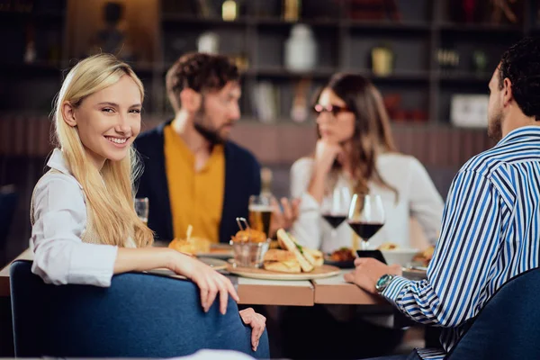 Fiatal, mosolygós, kaukázusi szőke, csinos nő elegáns alkalmi ruhában ül az asztalnál az étteremben, és a válla fölé néz. A háttérben a barátai vacsoráznak.. — Stock Fotó