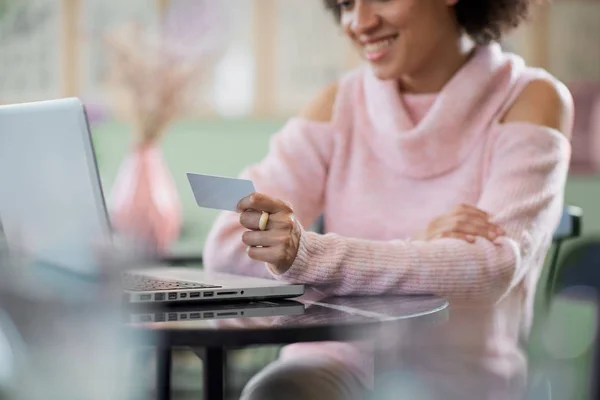 Vacker blandad ras kvinna i rosa polotröja sitter i konditoriet och använder laptop för on line shopping. — Stockfoto