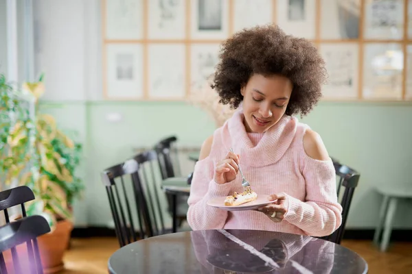 Portret van charmante positieve mixed race vrouw in roze coltrui zitten in zoetwaren en eten gateau. — Stockfoto