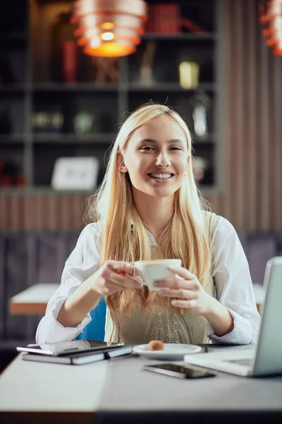 Charmante lachende blonde zakenvrouw gekleed Smart Casual zitten in cafetaria, koffie drinken en laptop gebruiken. — Stockfoto
