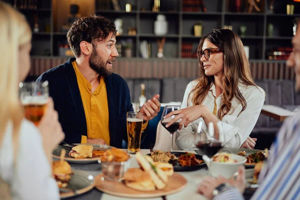 Amigos multiétnicos sentados no restaurante, bebendo álcool, conversando e comendo hambúrgueres para o jantar . — Fotografia de Stock