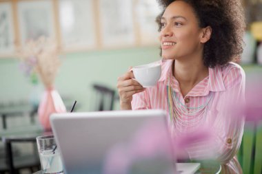 Kahve dükkanında aitting çizgili pembe elbise büyüleyici karışık yarış kadın, kahve içme ve dizüstü bilgisayar kullanarak.