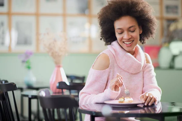 Portret van charmante positieve mixed race vrouw in roze coltrui zitten in zoetwaren en eten gateau. — Stockfoto