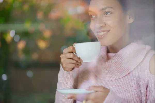 Gorgeous mixed race woman in pink turtleneck sweater standing in coffee shop, drinking espresso and looking trough window. — Stock Photo, Image