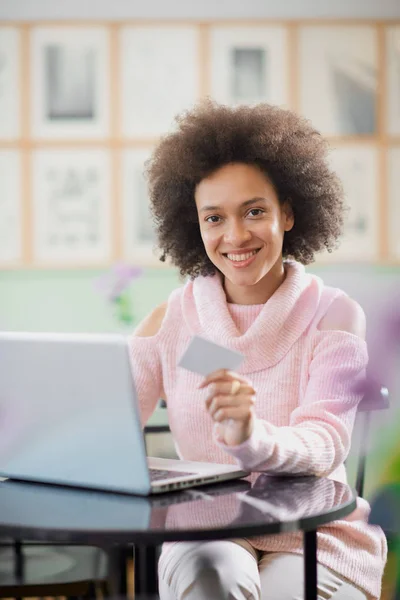 Vacker blandad ras kvinna i rosa polotröja sitter i konditoriet och använder laptop för on line shopping. — Stockfoto