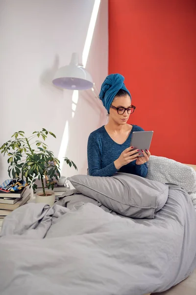 Hermosa morena con anteojos y toalla en la cabeza usando tableta mientras está sentada en la cama en el dormitorio . — Foto de Stock