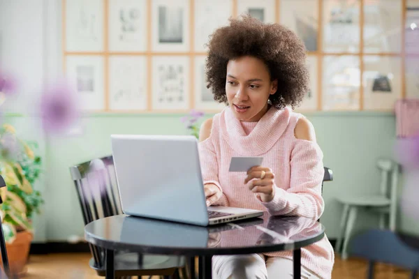 Hermosa mujer de raza mixta en suéter de cuello alto rosa sentado en la pastelería y el uso de ordenador portátil para ir de compras en línea . — Foto de Stock
