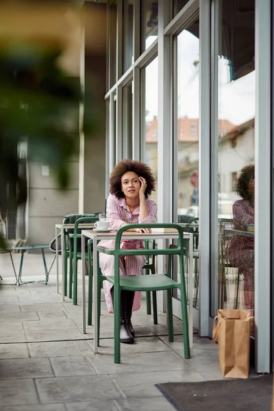 Belle femme métissée aux cheveux bouclés et vêtue d'une robe à rayures roses s'appuyant sur la table et la pensée. Café extérieur . — Photo