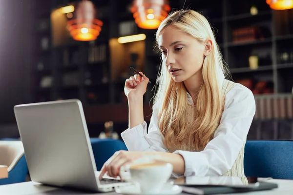 Jovem mulher de negócios loira caucasiana vestida casual inteligente usando laptop e segurando caneta enquanto sentada no café . — Fotografia de Stock