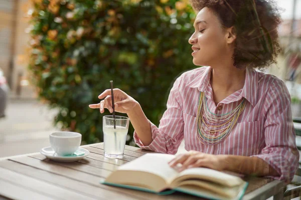 Linda mujer de raza mixta en vestido de rayas rosa sentado en un café, beber limonada y leer libro . — Foto de Stock