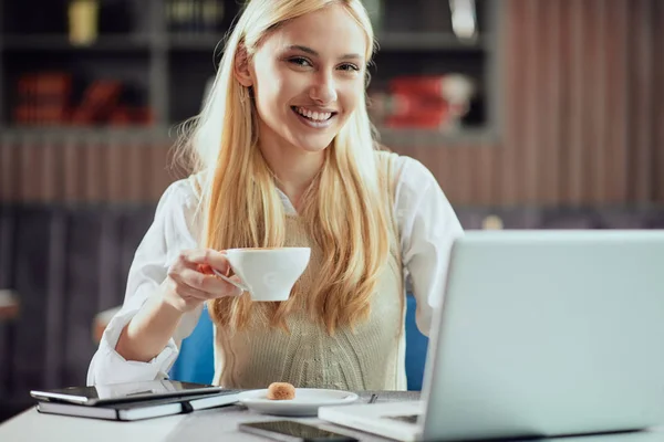 Encantadora mulher de negócios loira sorridente vestida inteligente casual sentado na cafetaria, bebendo café e usando laptop . — Fotografia de Stock