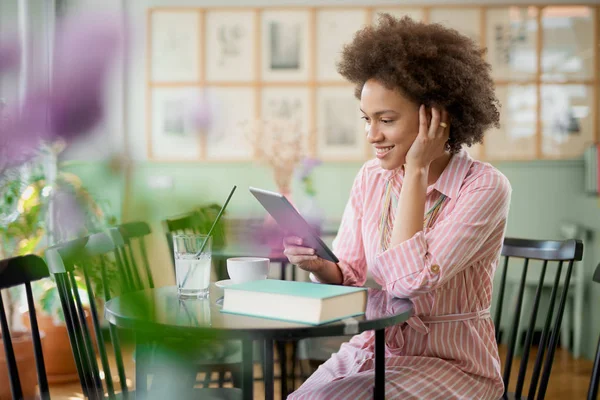 Attraktiv blandad ras kvinna i randig rosa klänning sitter i kafé och använder Tablet. — Stockfoto