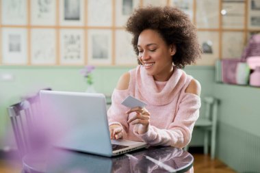 Pasta dükkanında oturan ve hat alışveriş için dizüstü bilgisayar kullanarak pembe balıkçı yakalı kazak güzel karışık yarış kadın.