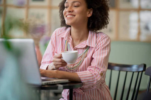 Charmante mixed race vrouw in gestreepte roze jurk aitting in Coffee Shop, koffie drinken en het gebruik van laptop. — Stockfoto