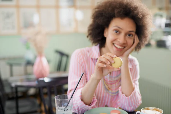 Schöne lächelnde Mischlingsfrau in rosa gestreiftem Kleid, die in der Konditorei sitzt und Kekse isst. — Stockfoto
