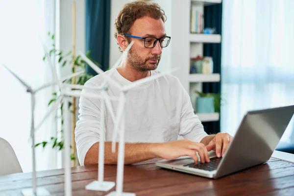 Ingeniero de ecología guapo sentado en casa, escribiendo en el ordenador portátil y trabajando en el proyecto. En el escritorio hay modelos de molino de viento. Concepto de desarrollo sostenible . — Foto de Stock