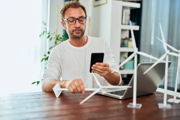 Ingeniero guapo de ecología caucásica sentado en casa y tomando fotos del modelo de molino de viento. En el escritorio hay modelos de molino de viento. Concepto de desarrollo sostenible . — Foto de Stock