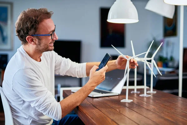 Knappe blanke ecologie ingenieur zittend thuis en het nemen van foto van windmolen model. Op het Bureau zijn windmolen modellen. Concept duurzame ontwikkeling. — Stockfoto