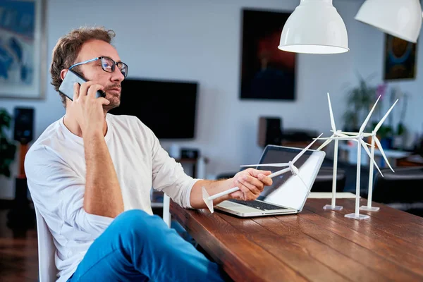 Knappe blanke ecologie ingenieur zittend thuis, praten over de telefoon over project en holding windmolen model. Op het Bureau zijn windmolen modellen. Concept duurzame ontwikkeling. — Stockfoto