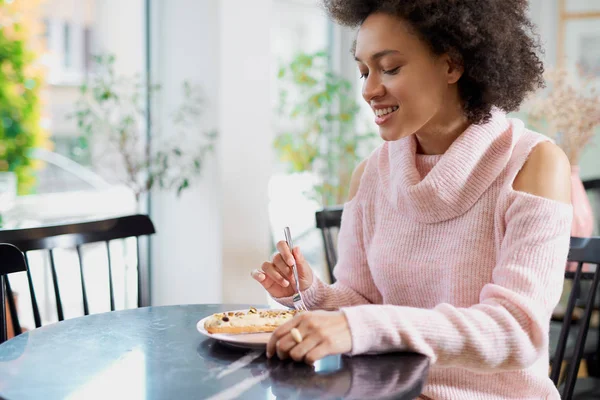 Portret van charmante positieve mixed race vrouw in roze coltrui zitten in zoetwaren en eten gateau. — Stockfoto