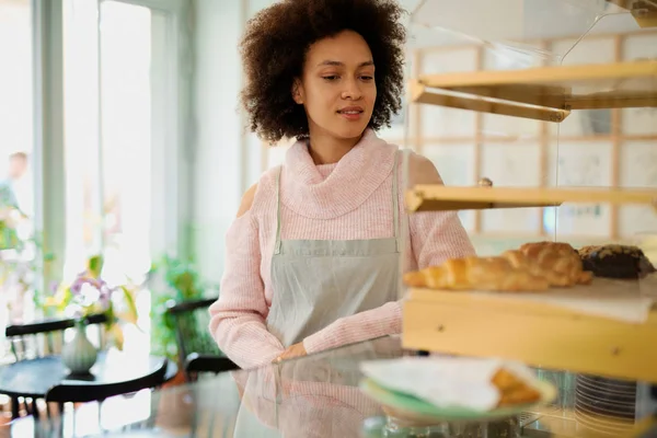 Beautiful smiling mixed race female employee in apron posing in pastry shop next to counter. — Stock Photo, Image