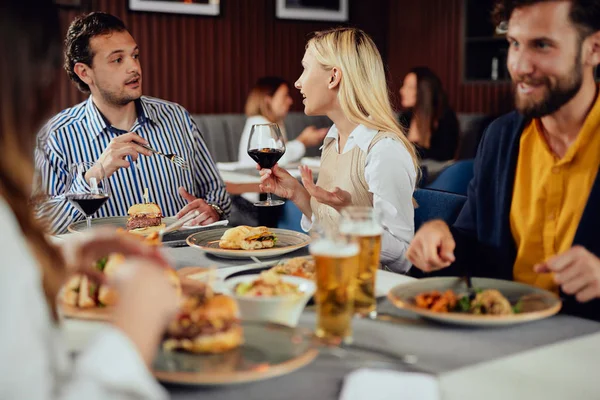 Multietnické přátele, kteří sedí v restauraci, konzumují alkohol, chatují a mají hamburgery na večeři. — Stock fotografie