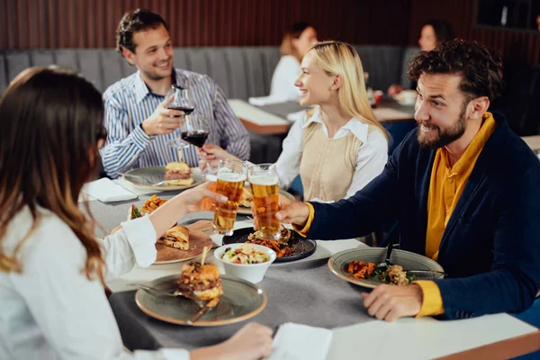 Multietnické přátele, kteří sedí v restauraci, konzumují alkohol, chatují a mají hamburgery na večeři. — Stock fotografie