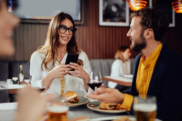 Multietnické přátele, kteří sedí v restauraci, konzumují alkohol, chatují a mají hamburgery na večeři. — Stock fotografie