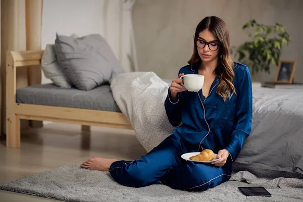 Młoda kaukaski brunetka w piżamie picie kawy, trzymając płytkę z rogalik i siedzi na podłodze w sypialni w godzinach porannych. — Zdjęcie stockowe