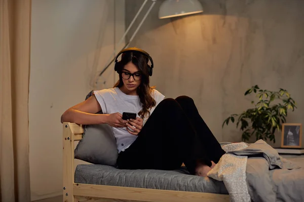 Pretty Caucasian brunette in pajamas and with headphones on head using smart phone late at night while sitting on the bed in bedroom. — Stock Photo, Image