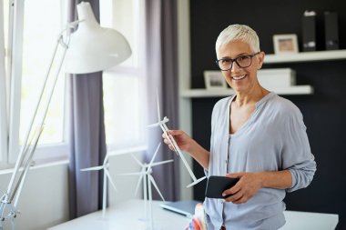 Güzel gülümseyen Kafkas yaşlı kadın ofiste ayakta, yel değirmeni modeli ve akıllı telefon tutan. İş konseptini başlatın.