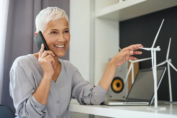 Великолепная белая старшая женщина разговаривает по телефону и трогает модель мельницы, сидя в офисе. Концепция стартапа . — стоковое фото