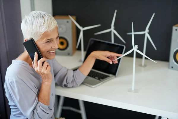 Preciosa mujer mayor caucásica hablando por teléfono y tocando el modelo de molino de viento mientras está sentado en la oficina. Concepto de creación de empresas . — Foto de Stock