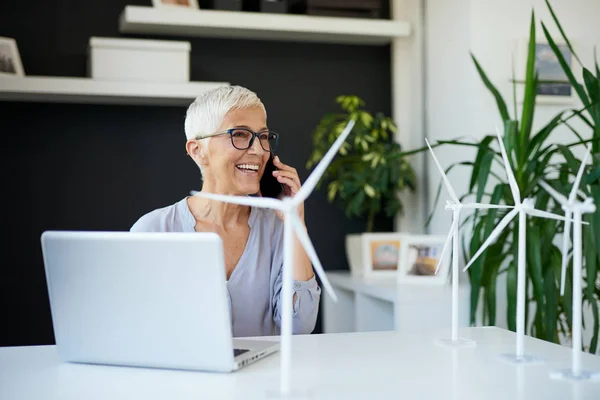 Великолепная белая старшая женщина разговаривает по телефону, сидя в офисе. На столе находятся модели ветряной мельницы и laptop.Start бизнес-концепции . — стоковое фото
