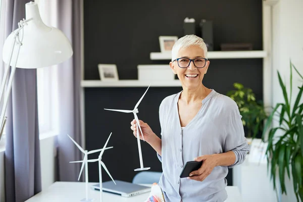 Красивая улыбающаяся белая старшая женщина, стоящая в офисе, держа в руках модель мельницы и смартфон. Концепция стартапа . — стоковое фото
